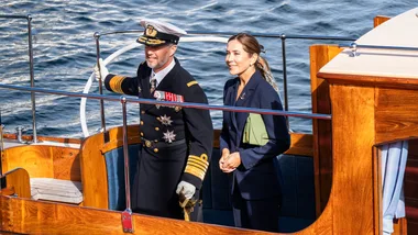 A man in a naval uniform and a woman in a suit on a wooden boat, with water in the background.