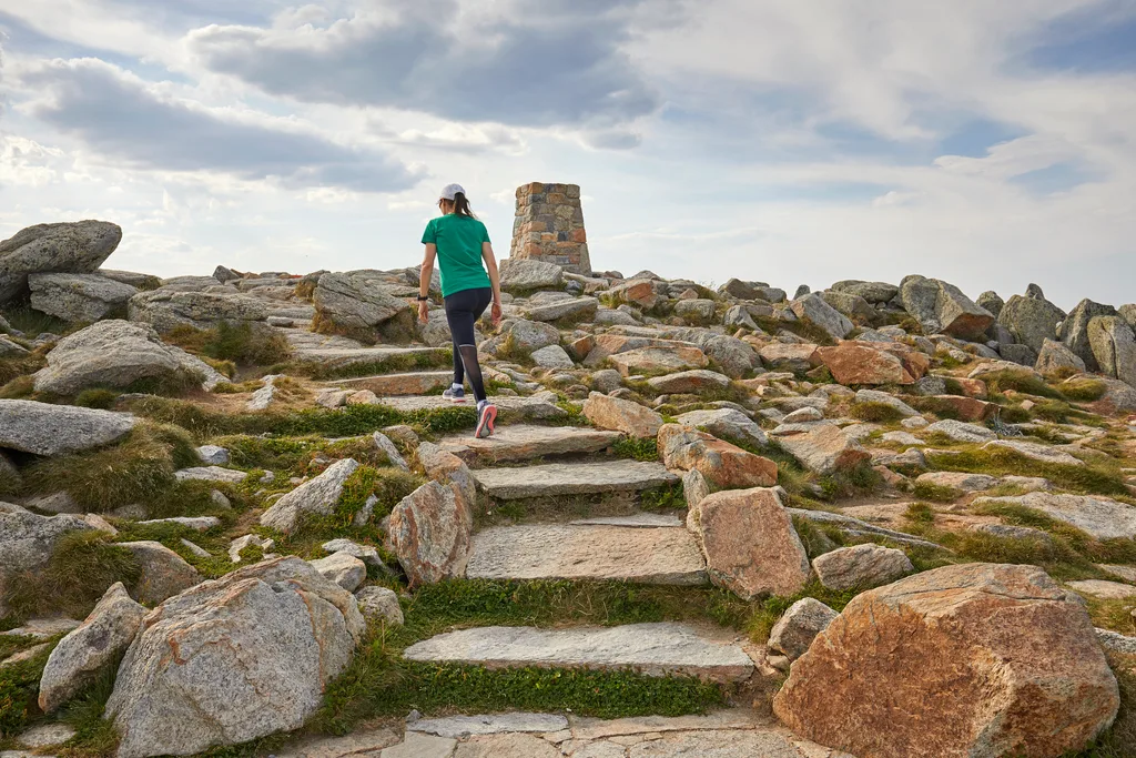 Person hiking up rocky steps toward a stone cairn under a cloudy sky.