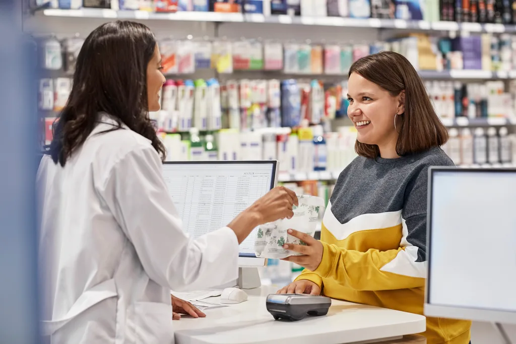 Pharmacist in white coat handing a prescription bag to a smiling customer at the pharmacy counter.