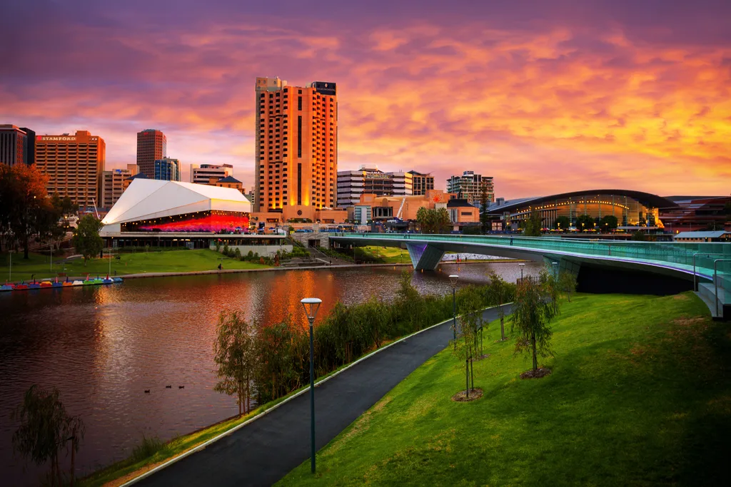 Adelaide skyline at sunset, featuring the Festival Centre, modern buildings, and a curved footbridge over the river.