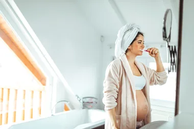 Pregnant woman brushing teeth in bathroom, wearing a bathrobe and towel on head.
