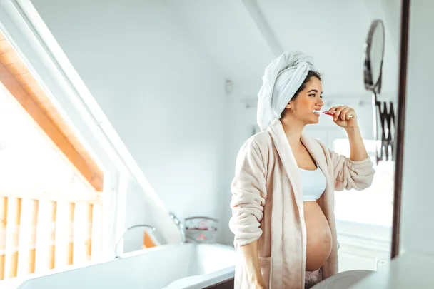 Pregnant woman brushing teeth in bathroom, wearing a bathrobe and towel on head.