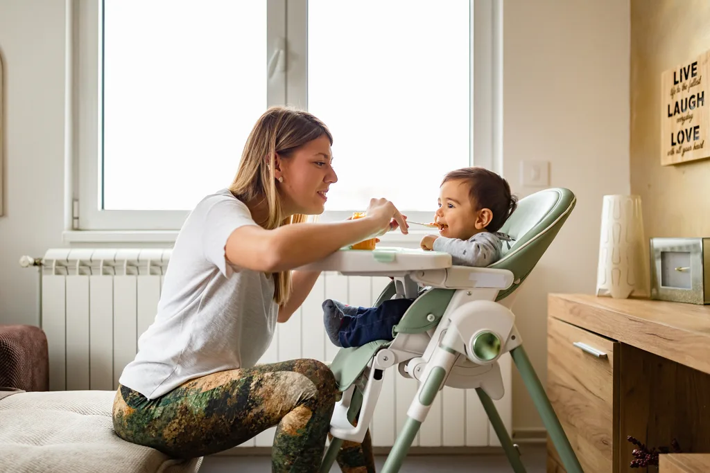 Mother feeding baby in a high chair near a window, with a motivational sign on the wall.