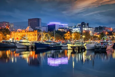 Boats docked at vibrant Hobart waterfront, illuminated buildings reflecting on calm water under a cloudy dusk sky.