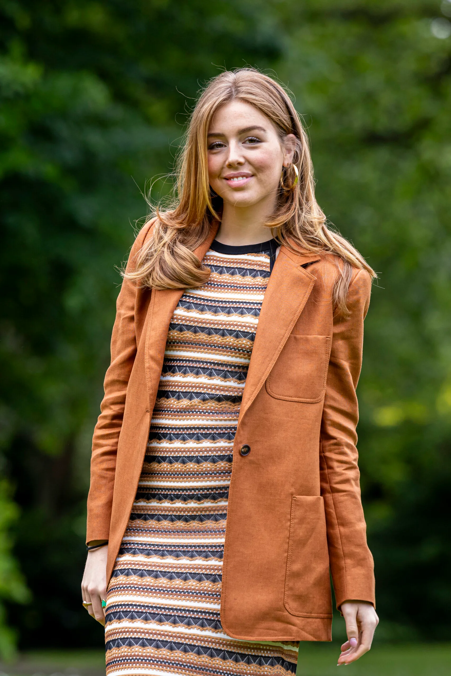 A young woman with light brown hair in an orange jacket and patterned dress, standing outdoors with a blurred green background.
