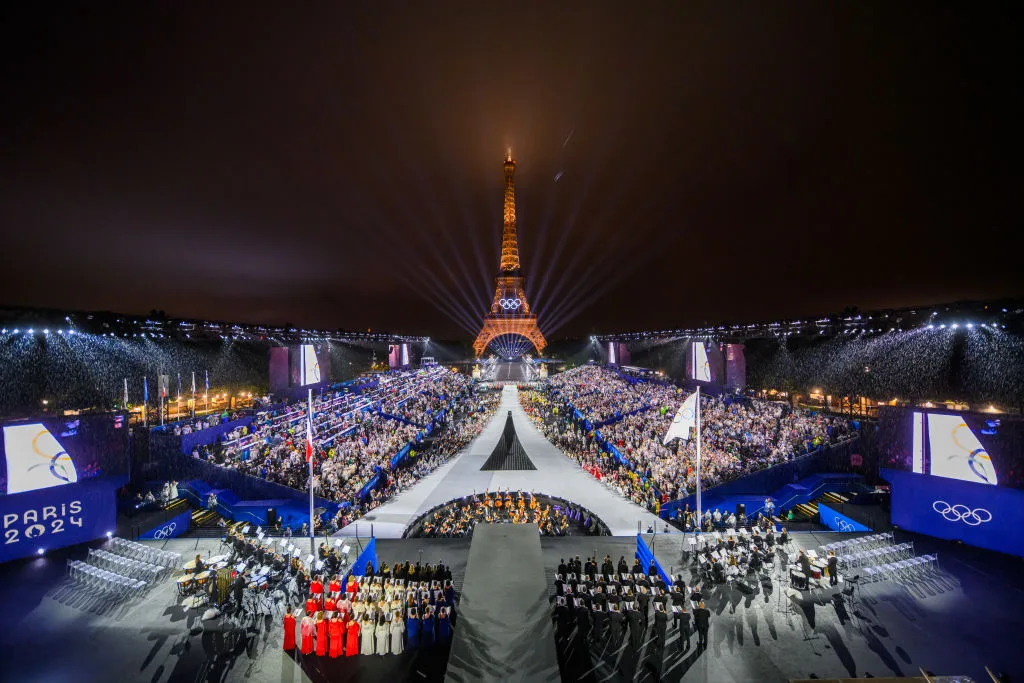 Paris 2024 Olympic ceremony beneath the Eiffel Tower, with large crowd and illuminated atmosphere at night.
