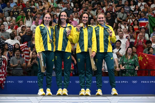 Australian women's team smiling with gold medals on Olympic podium, Paris 2024, with cheering crowd in background.
