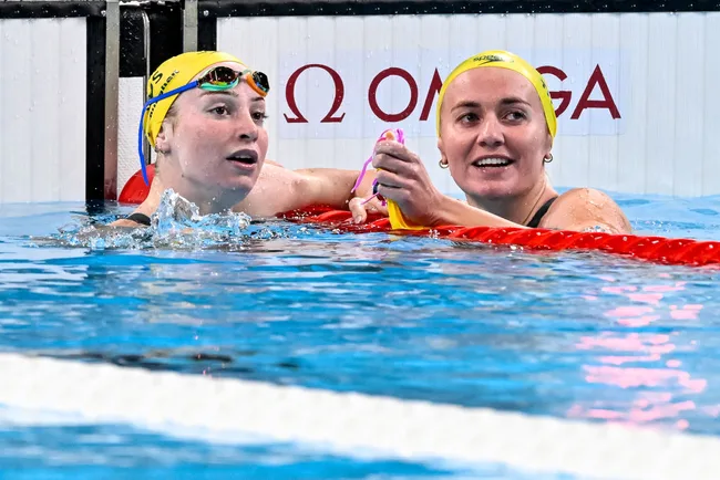 Two swimmers in yellow caps rest at the pool edge, smiling and talking after a race.