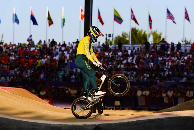 A cyclist in an Australian uniform performs a stunt at a BMX event with flags and a crowd in the background.