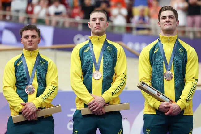 Three Australian cyclists in team uniforms on the podium with medals and awards at a cycling event.