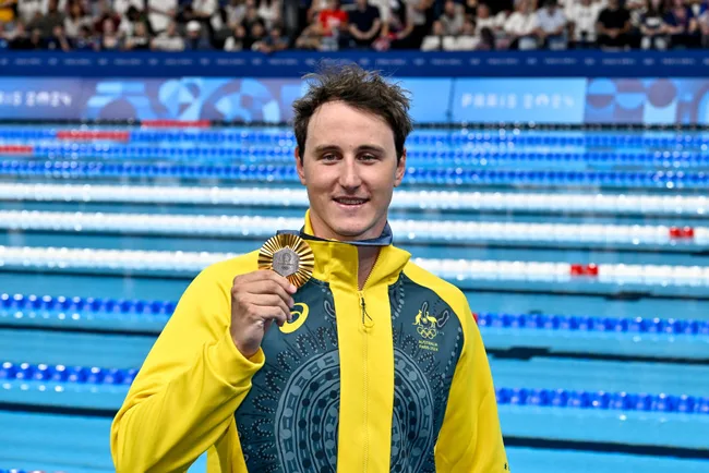 A male swimmer in a yellow jacket holds a medal, standing by the pool at a competition, with a cheering crowd in the background.