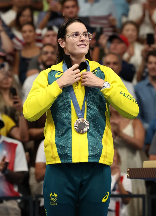 Australian athlete proudly holds a silver medal at a public event, wearing a yellow and green tracksuit, surrounded by a crowd.