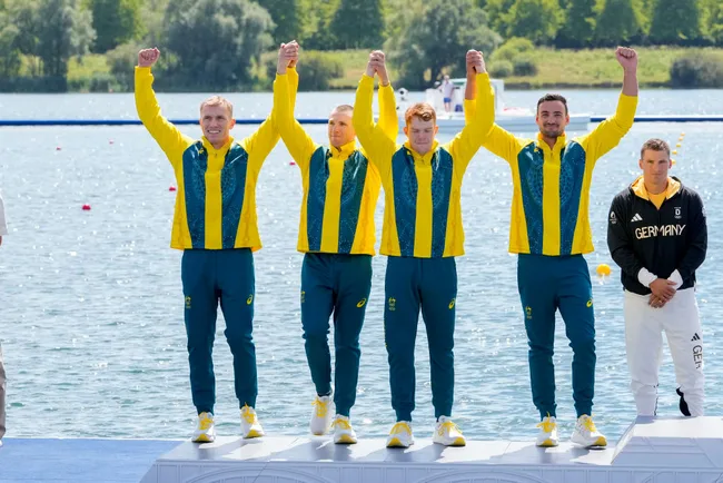 Australian rowing team celebrating on the podium at a lake, wearing yellow and green tracksuits, with German team nearby.