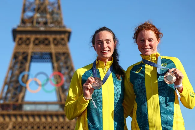 Two athletes in Australian uniforms holding medals, standing in front of the Eiffel Tower with Olympic rings.