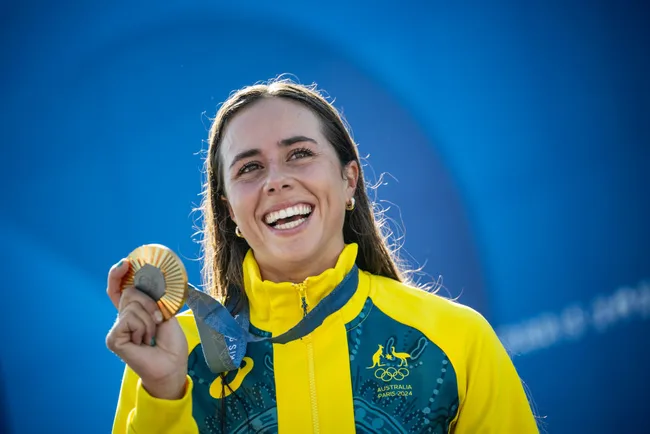 Smiling athlete in a yellow jacket holds a medal at the Paris 2024 event.