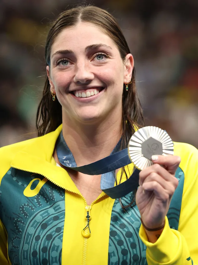 Smiling athlete wearing a yellow and green jacket holds a silver medal at an awards ceremony.