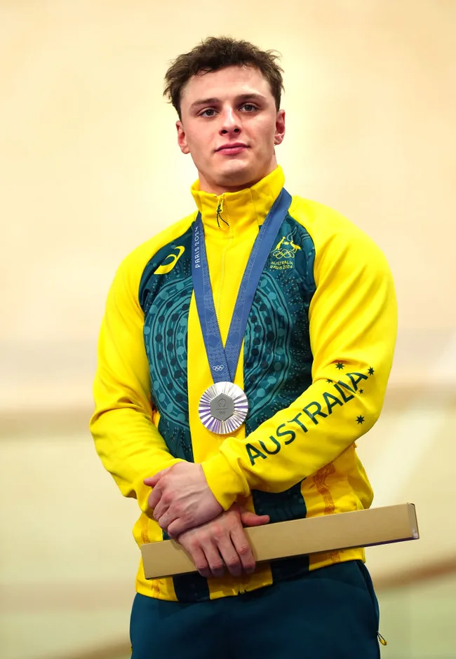 Australian athlete in a yellow jacket, wearing a silver medal, holding a rectangular box, standing proud against a light background.
