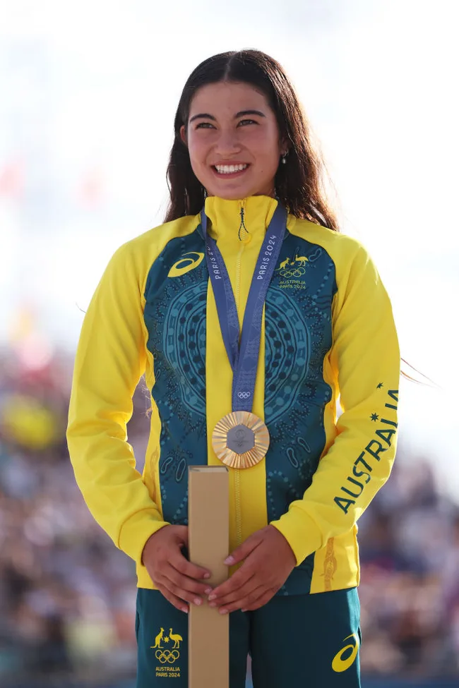 Australian athlete smiling with gold medal at Paris 2024 event, wearing a yellow and green jacket.