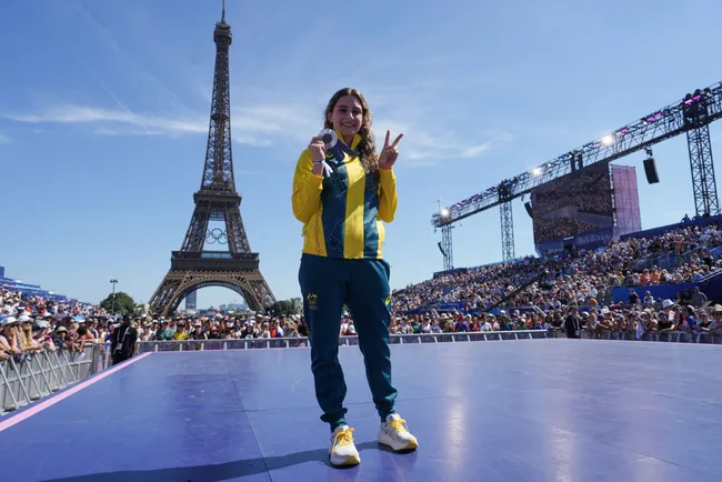 Female athlete holds medal and poses in front of Eiffel Tower, with crowd and stage, on a sunny day.