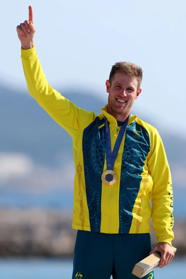 Athlete in a yellow and blue jacket with a medal, smiling and raising an arm, holding a box with blue sky background.