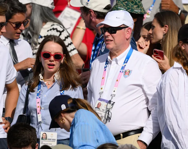 Spectators at a sports event wearing badges and sunglasses, surrounded by a crowd, outdoors on a sunny day.