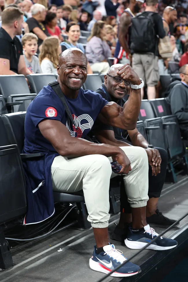 Two men smiling and flexing in a crowded arena, one wearing a navy shirt with a white and red design.