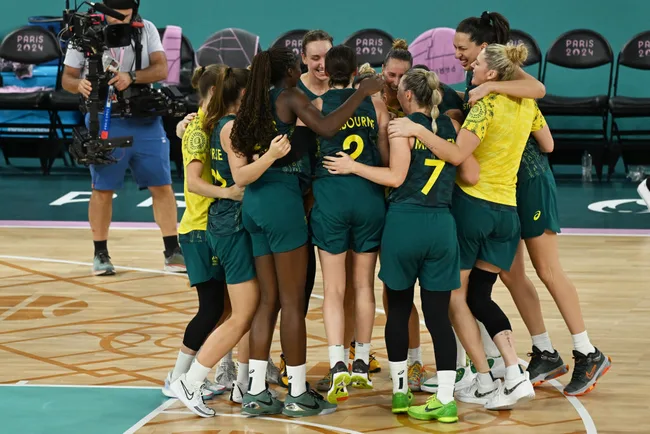 Australian women's basketball team huddles on court, wearing green and yellow uniforms, with Paris 2024 chairs in background.