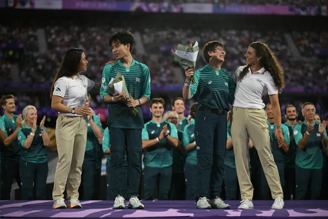 Two young athletes receive flowers on stage, surrounded by clapping people in sports uniforms at an event.