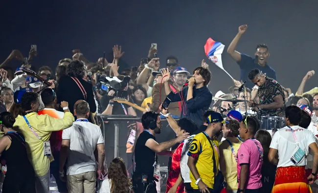 Lead singer performing amid a cheering crowd with a drummer and fans holding flags at a lively outdoor concert.