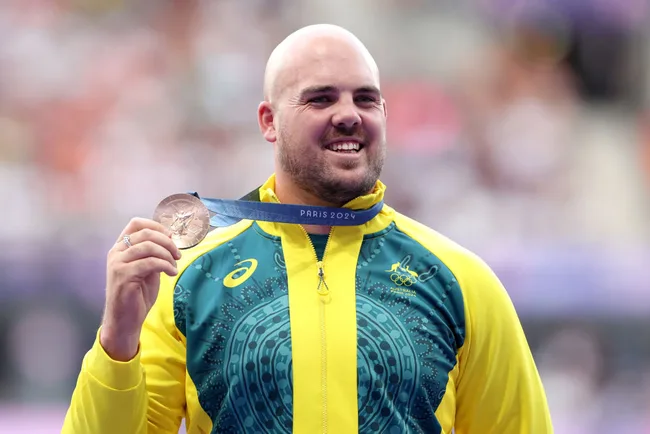 Athlete in a yellow and green jacket smiling and holding a bronze medal with "Paris 2024" written on the ribbon.