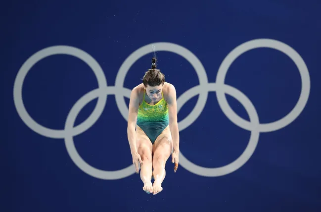 Diver in mid-air with Olympic rings in the background, wearing a green and yellow swimsuit.