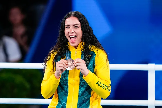 Australian female athlete wearing gold medal jacket, smiling and holding a bronze medal in front of a blue background.