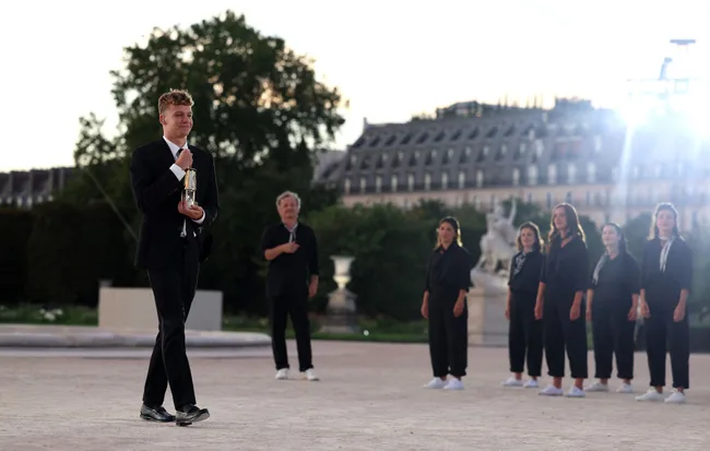 A man in a suit walks with an award, while a group of people stands in the background at a formal outdoor event.