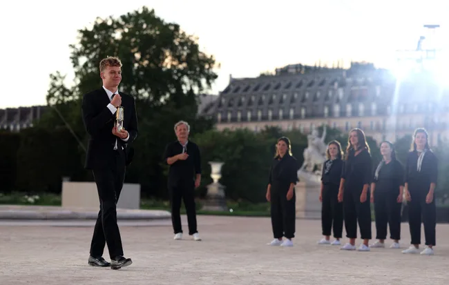 A man in a suit walks with an award, while a group of people stands in the background at a formal outdoor event.