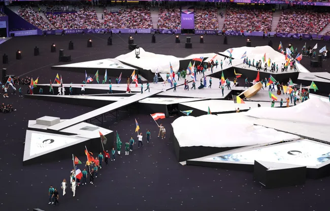 Athletes with flags walk on a geometric stage at the Paris 2024 Olympic opening ceremony, with a crowd in the background.