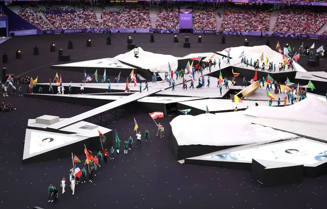 Athletes with flags walk on a geometric stage at the Paris 2024 Olympic opening ceremony, with a crowd in the background.