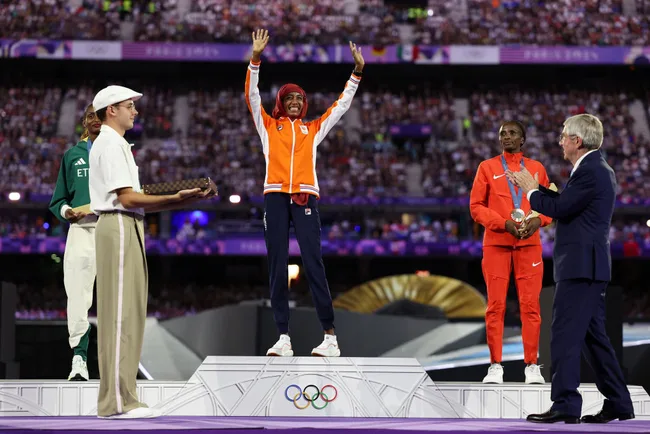 Athletes on Olympic podium; gold medalist celebrates with arms raised as medals are awarded to others on stage.