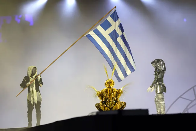 Three performers in costumes on stage; one holds a Greek flag, surrounded by fog and stage lights.