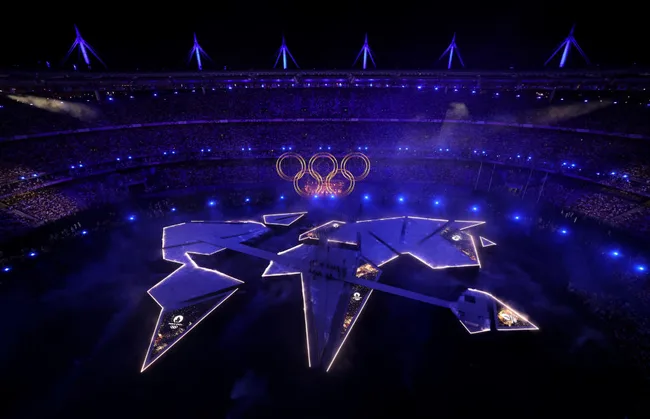 Aerial view of an illuminated stadium with Olympic rings during a night ceremony, featuring a star-shaped stage.