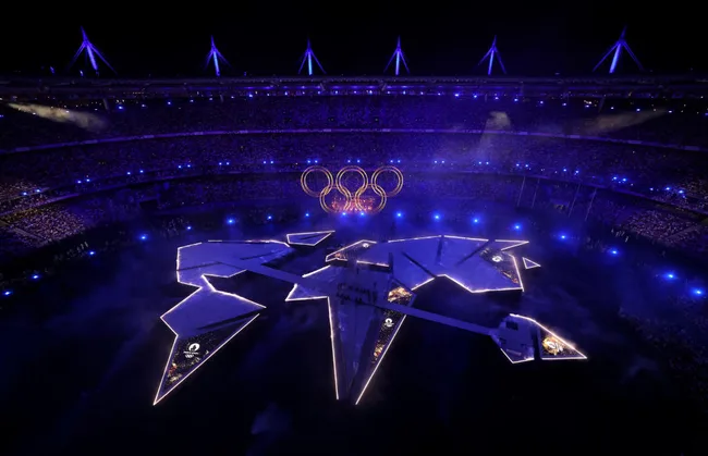 Aerial view of an illuminated stadium with Olympic rings during a night ceremony, featuring a star-shaped stage.