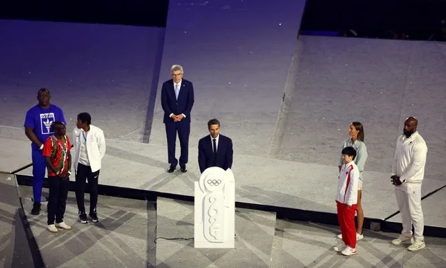 Seven people stand on a stage at an Olympic event, with a podium bearing the 2024 logo in front of them.