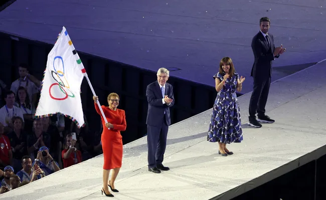 A woman waves an Olympic flag on stage, joined by two applauding individuals in formal attire, during a ceremony.