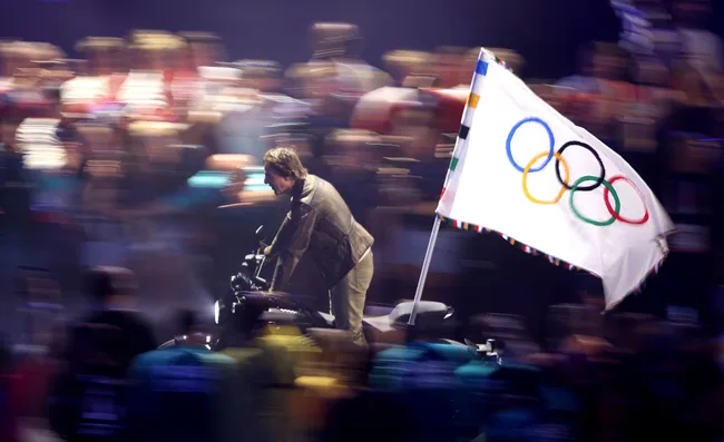 A man riding a motorcycle carries the Olympic flag, surrounded by a blurred crowd.