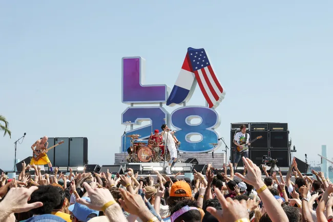 Band performing on stage with crowd in front, large "LA28" Olympic sign and USA flag, sunny outdoor event.