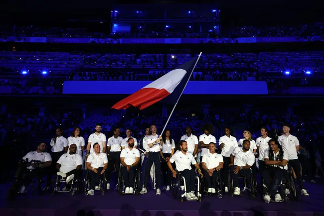 French athletes in white Polo shirts, some in wheelchairs, at a ceremony with a large French flag, under blue lighting.