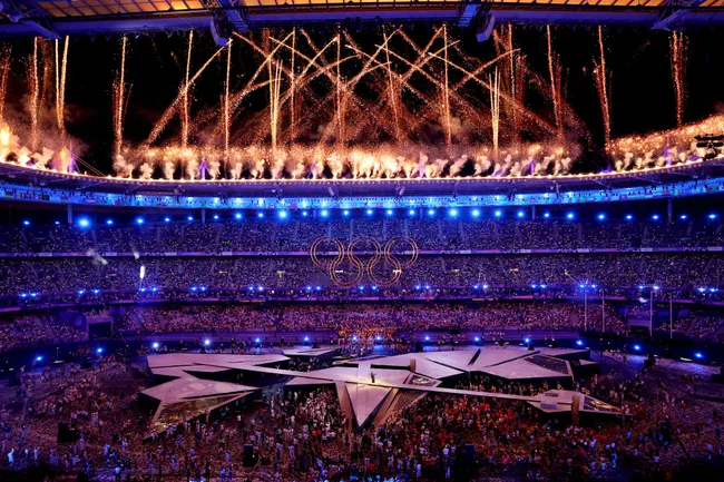Olympic stadium with vibrant fireworks and glowing rings during the opening ceremony, crowded with spectators and performers.
