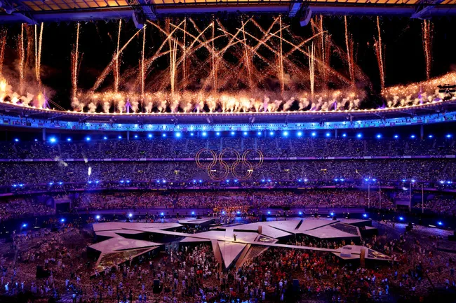 Olympic stadium with vibrant fireworks and glowing rings during the opening ceremony, crowded with spectators and performers.
