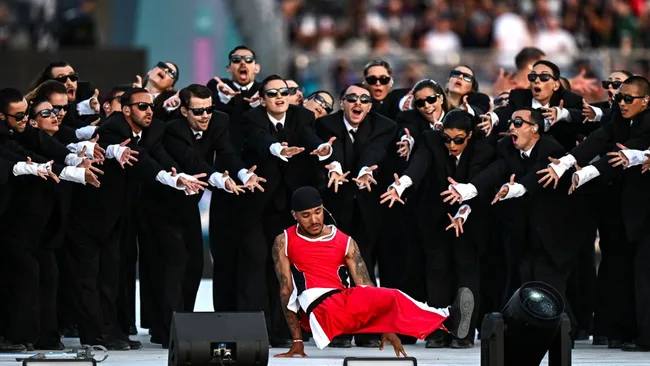 Performer in red costume dances in front of a group in black suits and sunglasses, reaching out dramatically.