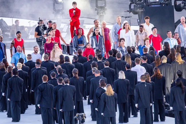 A large group of performers in colorful costumes on stage, with a crowd in dark suits watching them, during an outdoor event.