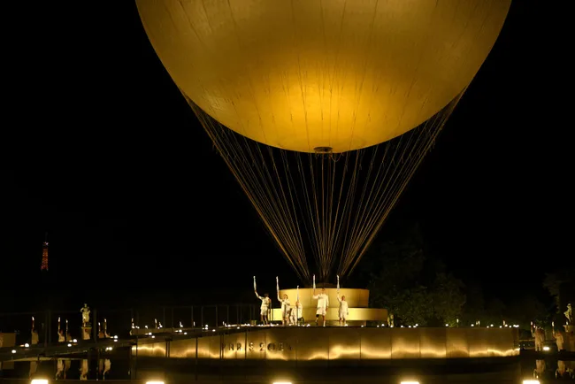 Night scene of Eiffel Tower and balloon installation at Paris 2024 Olympic Games event, with figures and lights beneath.